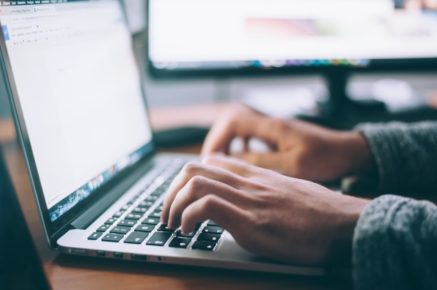 Hands typing on a keyboard with two monitors blurred in the background