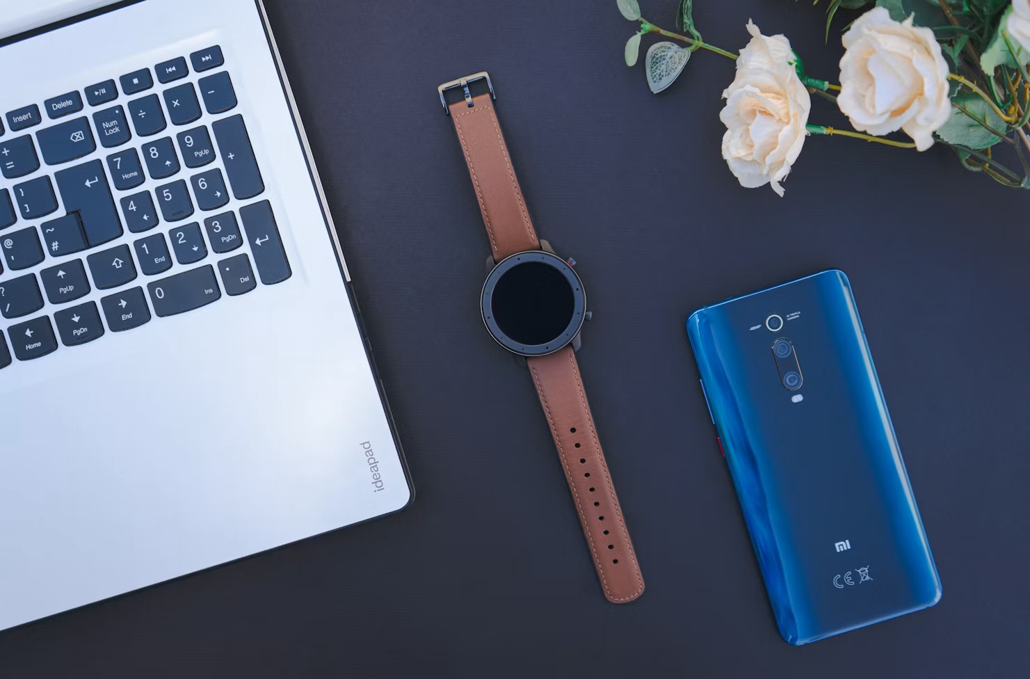A flat lay of a smartwatch, phone and laptop scattered with dried roses